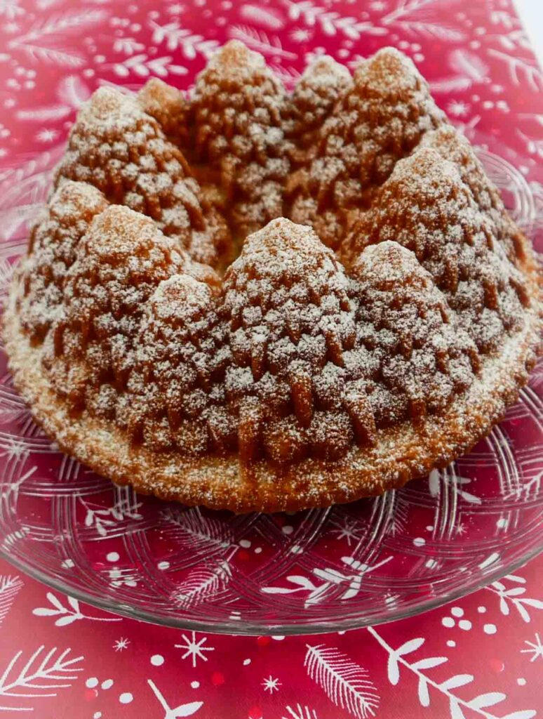 A Christmas Bundt cake in the shape of a circle of pine trees, dusted with icing sugar looking like snow. The cake is on a decorative glass plate on a red Christmas tablecloth.