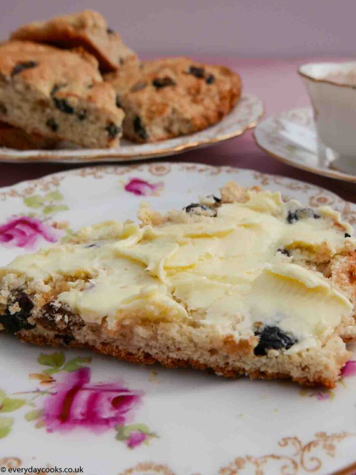 A slice of buttered fruit tea scone with a plate of scone a nd a cup of tea in the background. All on a pink tablecloth and on old-fashioned china with pink roses and a gold border.