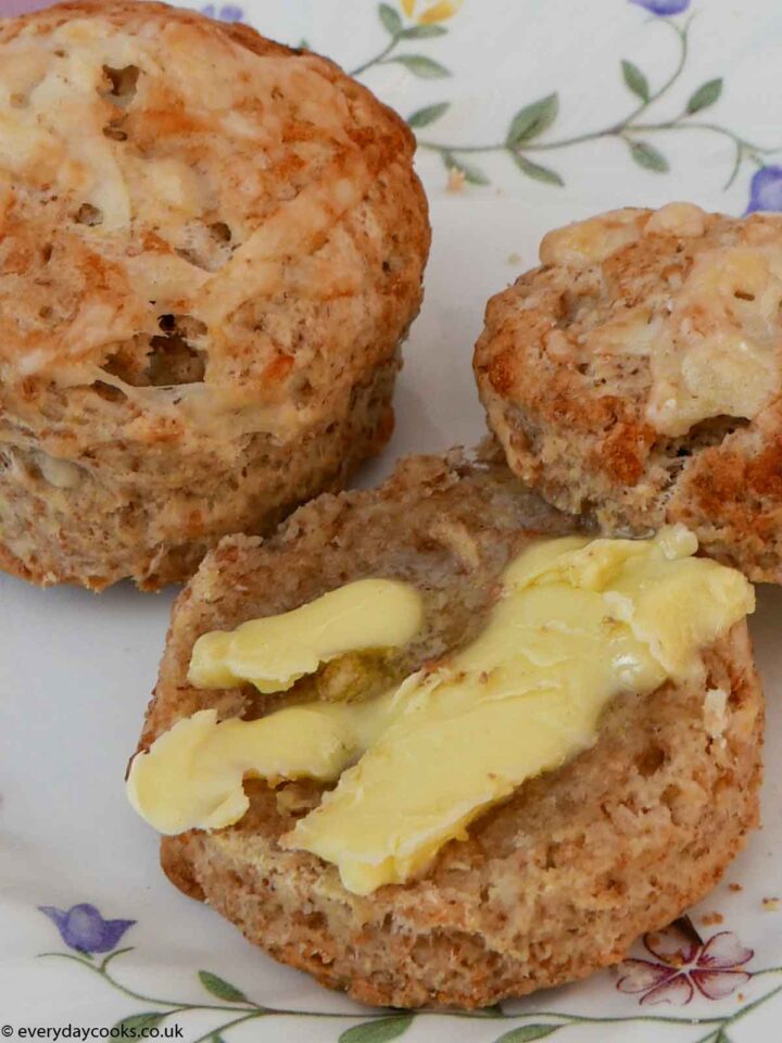 Two cheese scones on a flowered plate on a pink tablecloth. One cheese scone is split in two with one half buttered. The other scone is whole.