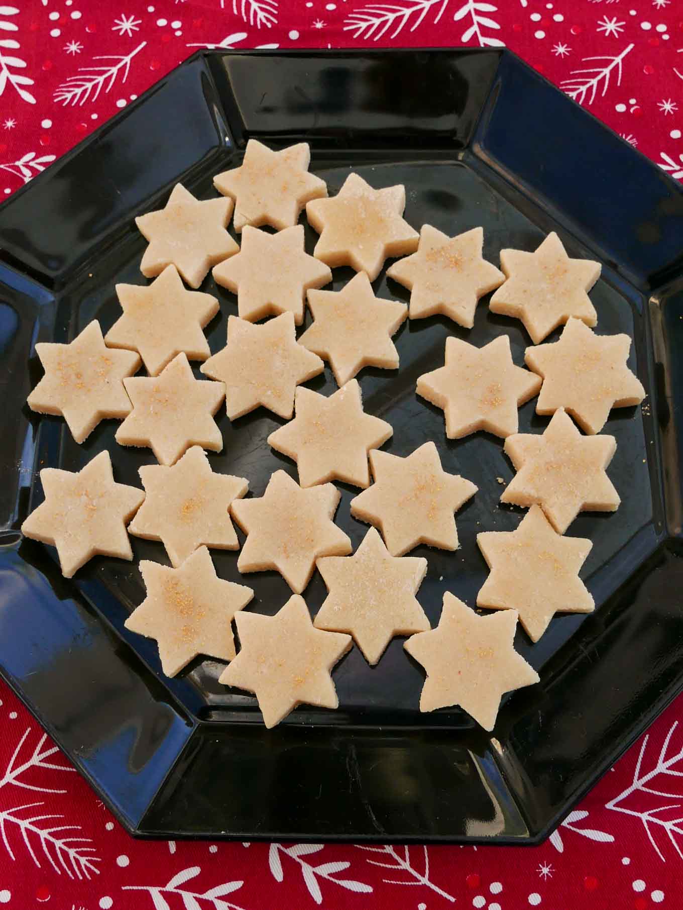 Marzipan Stars dusted with gold glitter and arranged on a black octagonal plate. It sits on a red Christmas table cloth