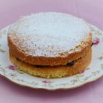 Victoria Sandwich cake with strawberry jam filling and icing sugar sprinkled on top. The cake is on a plate patterned with pink flowers and leaves.