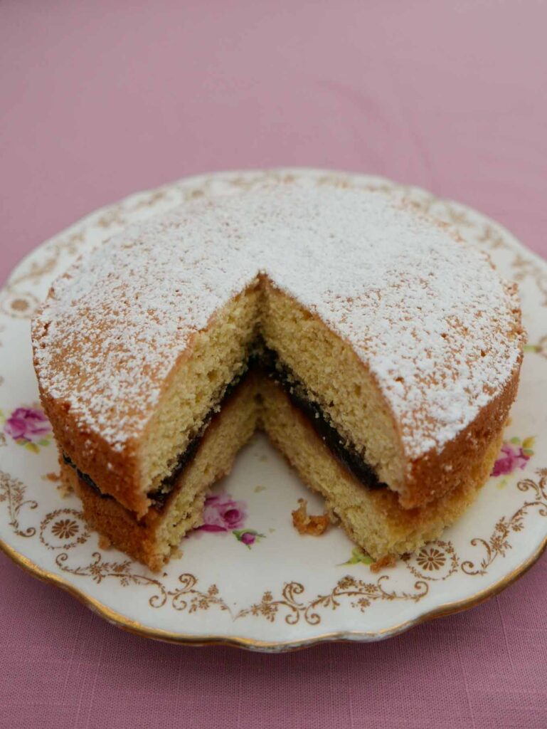 Victoria Sandwich cake with strawberry jam filling and icing sugar sprinkled on top. The cake is on a plate patterned with pink flowers and leaves.