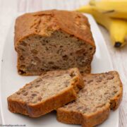Two slices of Banana Loaf on a white plate with the rest of the cake and 2 bananas in the background