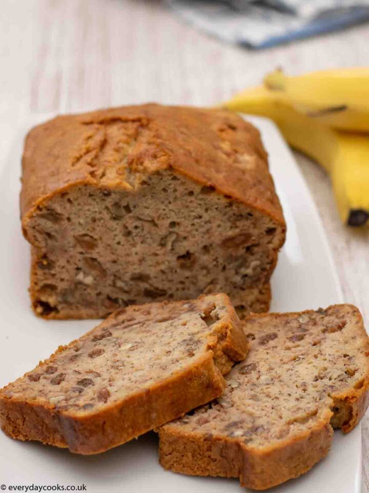 Two slices of Banana Loaf on a white plate with the rest of the cake and 2 bananas in the background