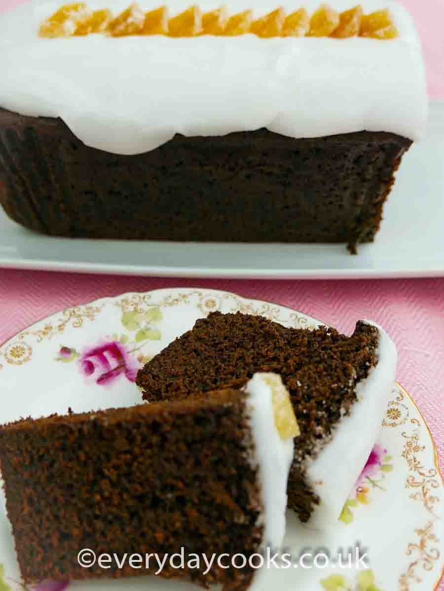 A slice of Iced Ginger Cake on a plate with the rest of the cake in the background