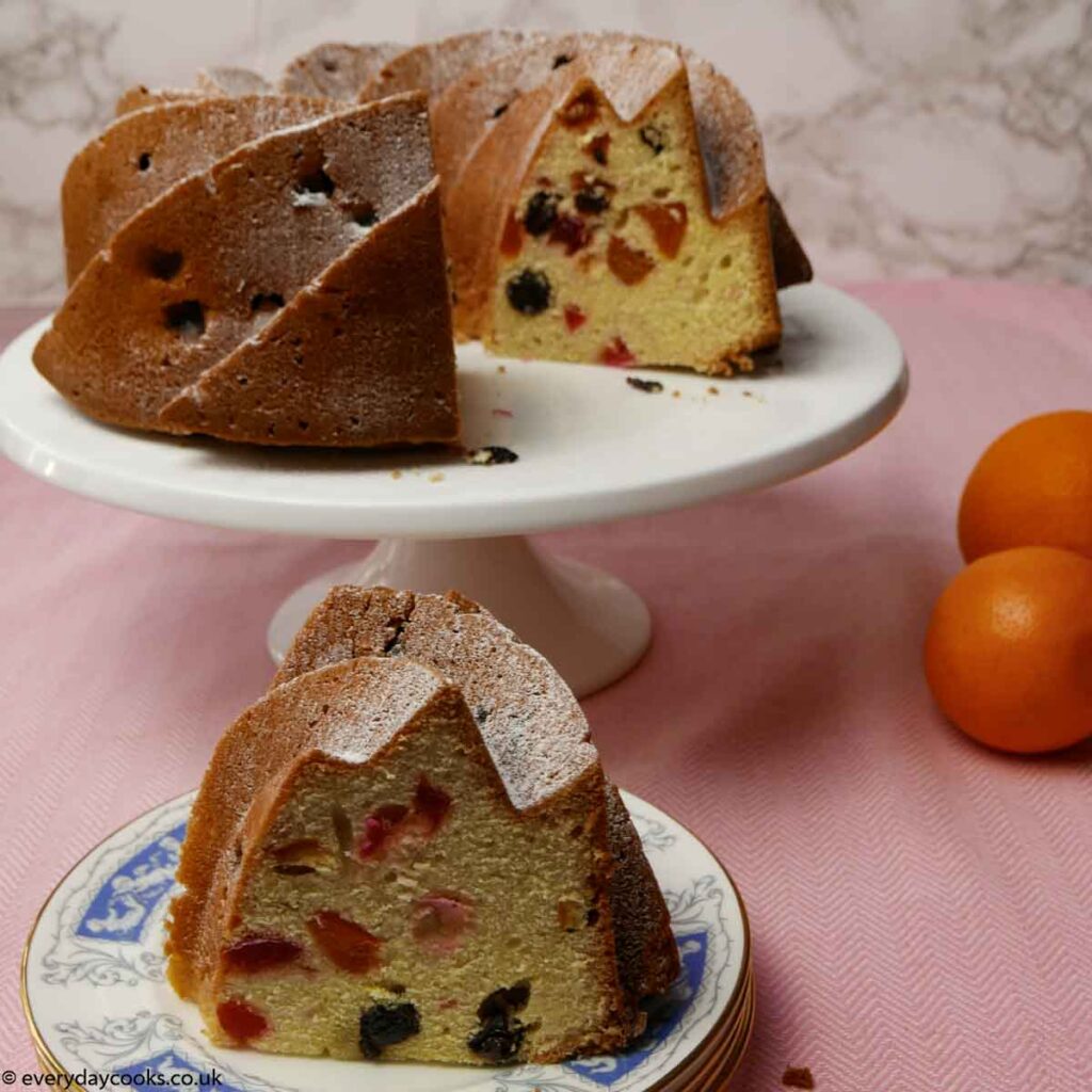 Fruit Bundt Cake on a white plate with a slice on a blue plate. With 3 oranges on a pink table cloth