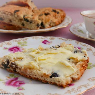 A slice of buttered fruit tea scone with a plate of scone a nd a cup of tea in the background. All on a pink tablecloth and on old-fashioned china with pink roses and a gold border.