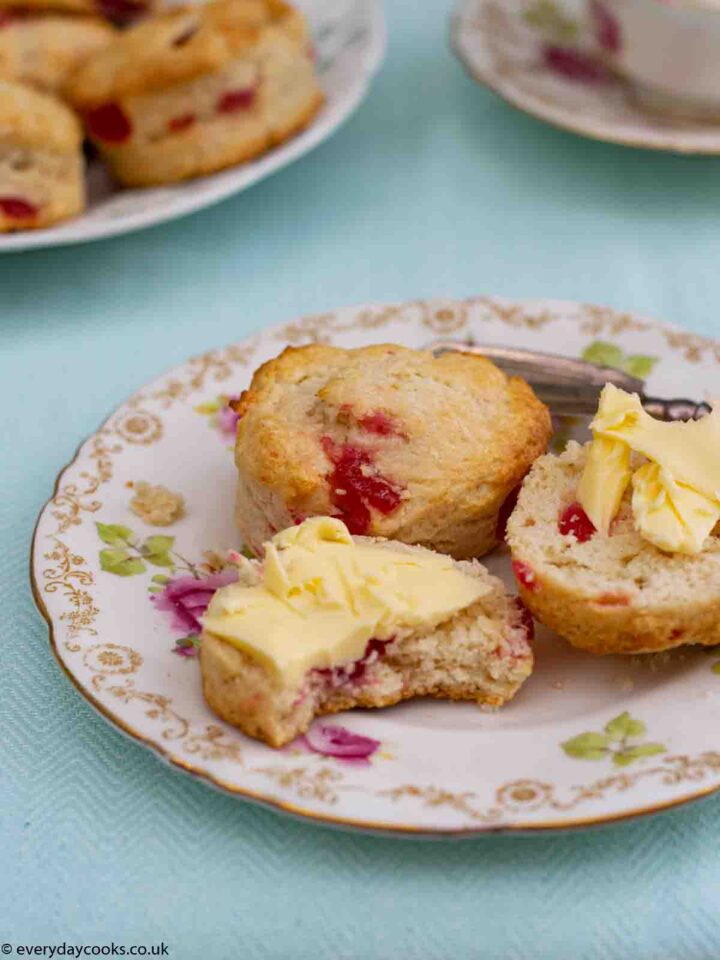 A Cherry Scone cut in half with butter and a whole scone on a patterned plate. More scones and a teacup int he background