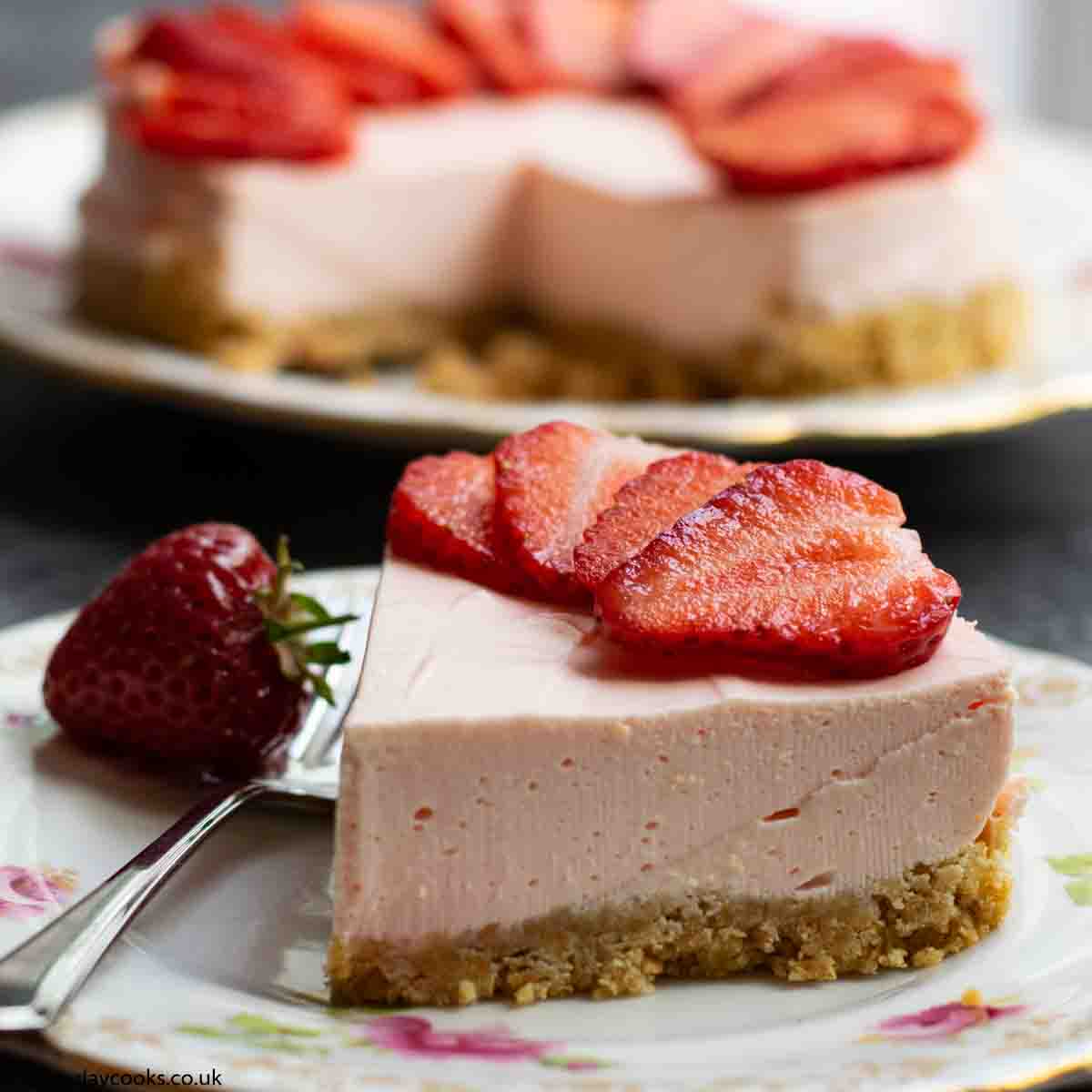 A slice of Easy Strawberry Cheesecake topped with sliced strawberries on a patterned plate on a black worktop. The rest of the cheesecake is in the background.