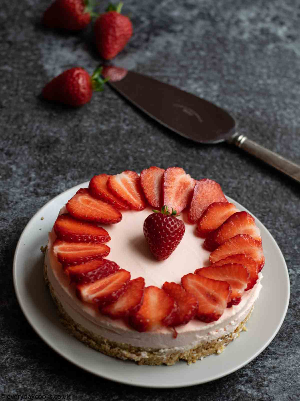 Easy Strawberry Cheesecake topped with sliced strawberries on a grey plate on a black worktop. 3 strawberries and a cake knife in the background.