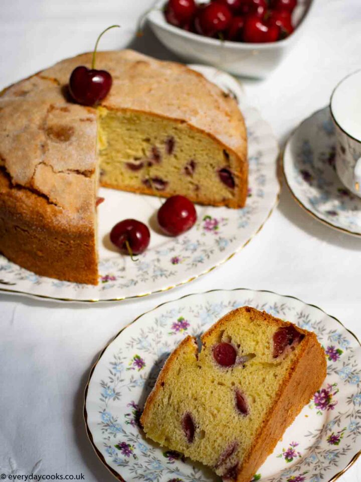 Fresh Cherry Cake, with a slice removed, on a patterned plate. A slice of cake on a small plate with a cup of tea and a bowl of cherries