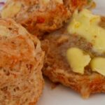 Two cheese scones on a flowered plate on a pink tablecloth. One cheese scone is split in two with one half buttered. The other scone is whole.