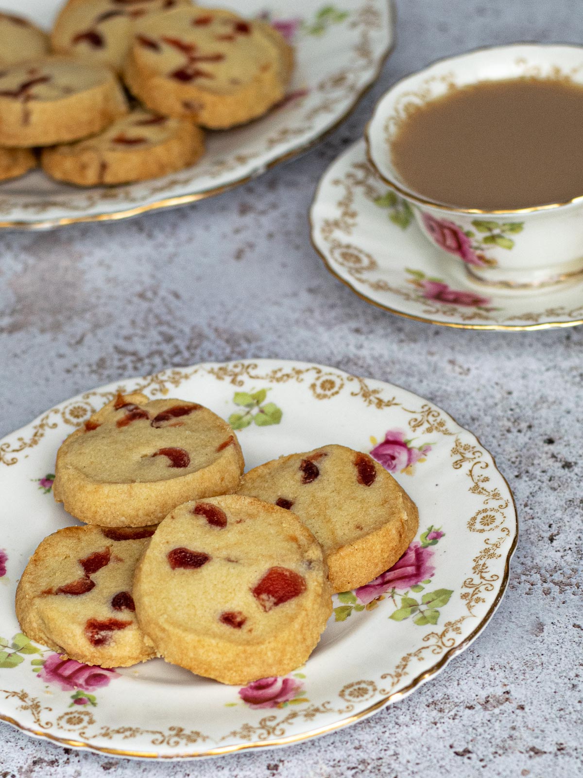 Four cherry shortbread biscuits on a pink and green patterned plate. A cup of tea and large plate of biscuits are in the background.