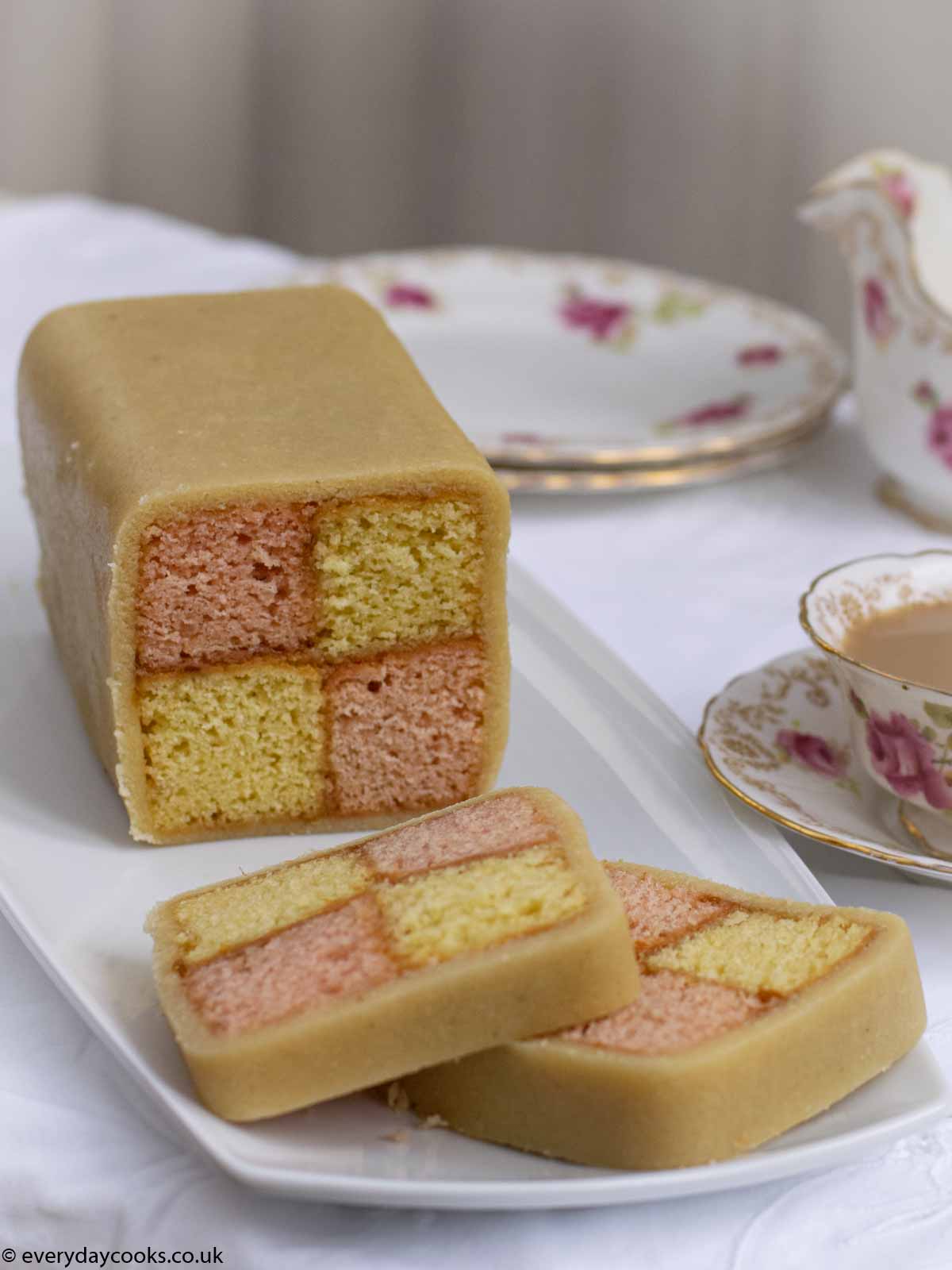 Easy Battenberg Cake on a white plate with 2 slices cut, with a cup of tea.