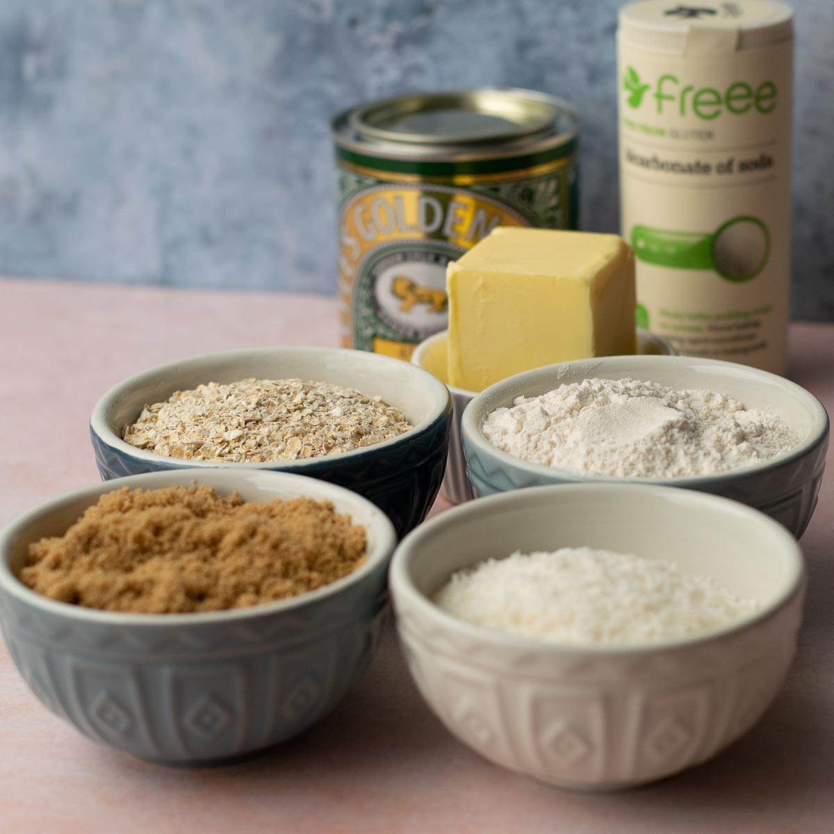 Blue and white bowls of ingredients for Anzac Biscuits: brown sugar, flour, coconut, oats, butter. Tin of golden syrup and pot of baking soda.