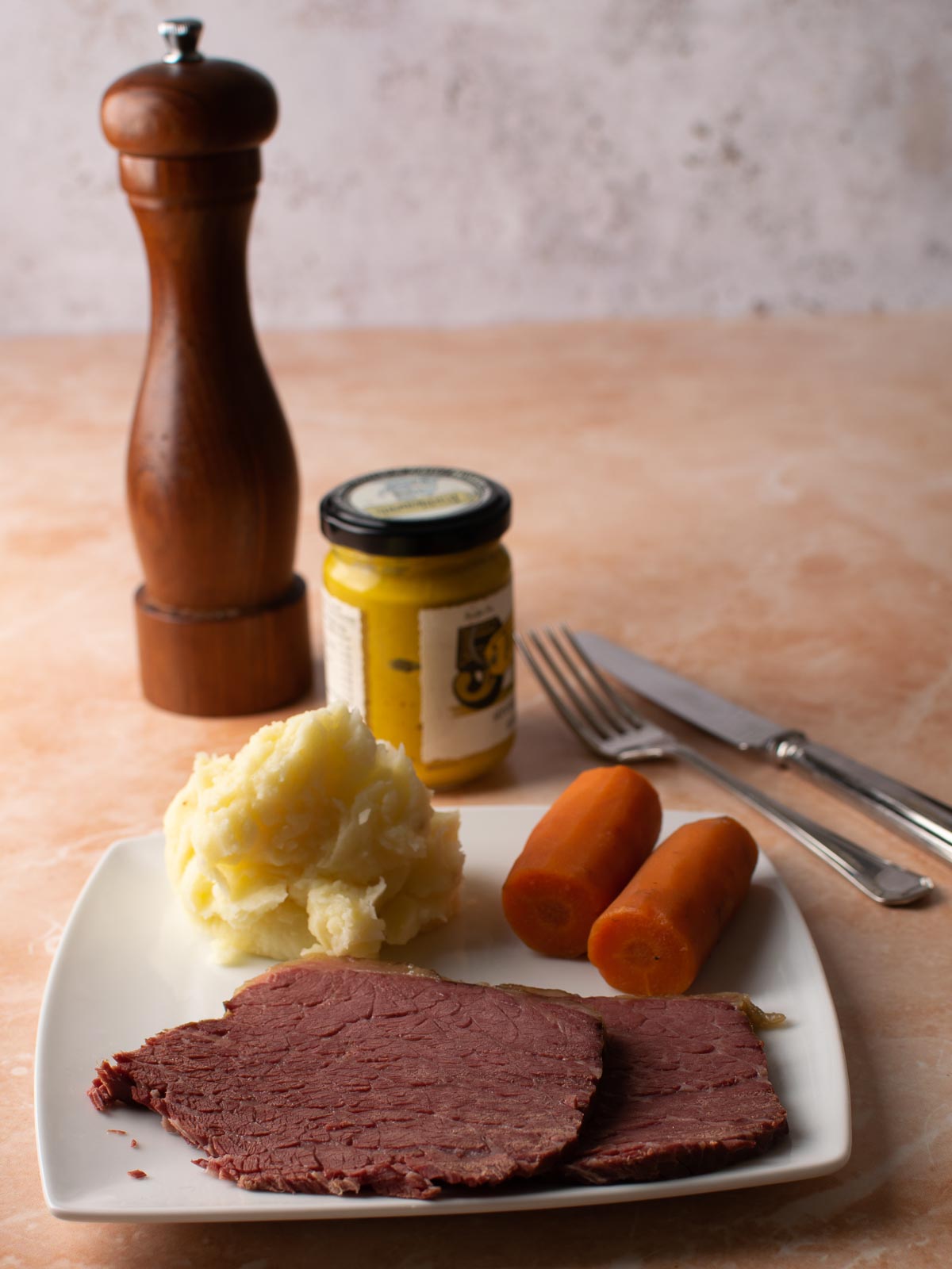 2 slices of Slow Cooker Salt Beef with carrots and mashed potato on a white plate. Jar of mustard, pepper ginder and cutlery in the background