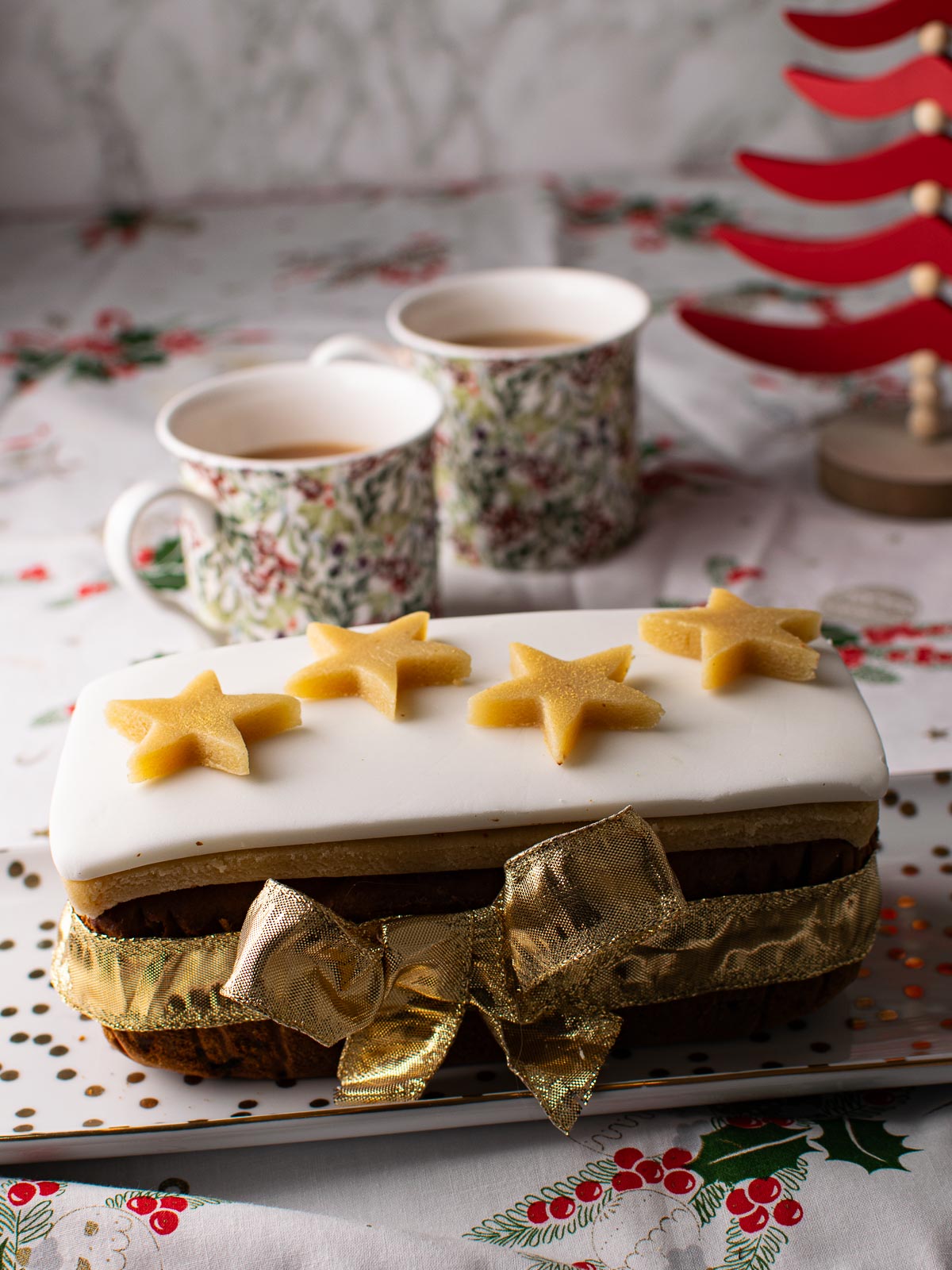 Small Christmas Loaf Cake decorated with marzipan, icing and marzipan stars. Two mugs of tea and a Christmas decorations in the background.