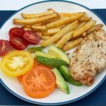 Air fried chicken and chips with avocado and red, yellow and orange tomatoes on a white plate with blue rim
